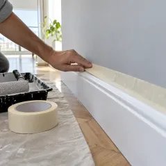 Mano pegando cinta de papel ancha sobre el borde de un zócalo blanco antes de pintar la pared