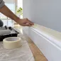 Mano pegando cinta de papel ancha sobre el borde de un zócalo blanco antes de pintar la pared