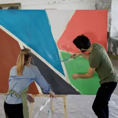 Dos personas pintando un mural geométrico y retirando la cinta de papel que enmascaró los bordes de cada color